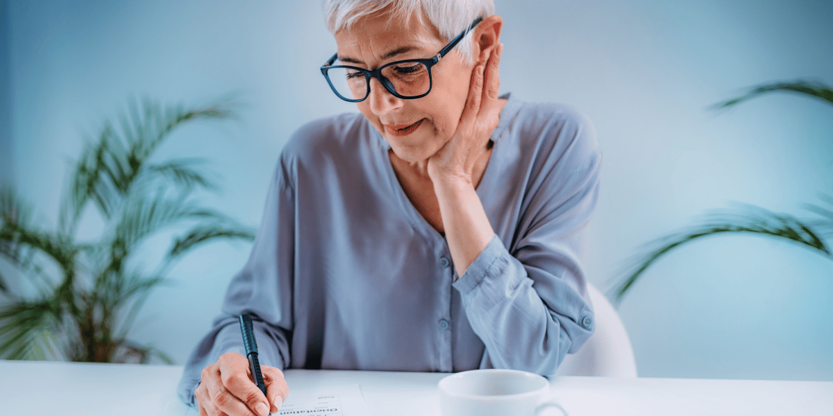 woman with gray hair and glasses thinking and writing on a table.